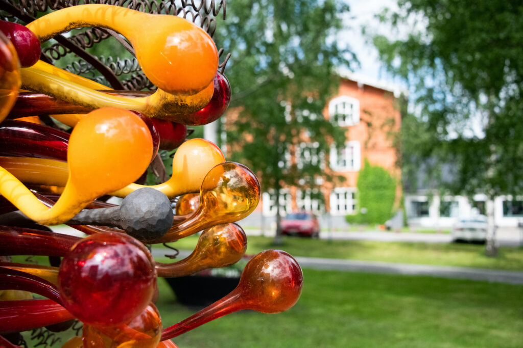 Värikas lasitaideteos etualalla, takana näkyy vanha punainen puutalo.