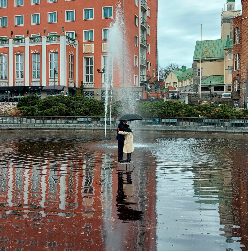 Kuva lammesta keskellä kaupunkia. Lammen keskellä seisoo pariskunta sateenvarjon alla.