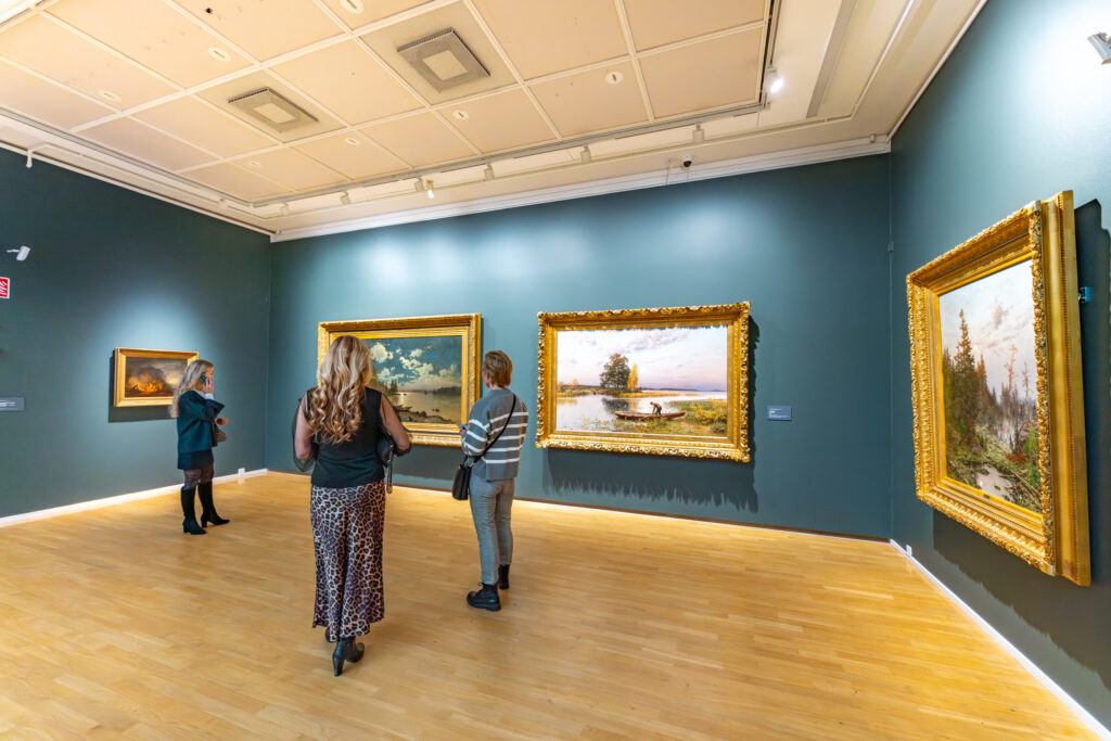 Siniseinäinen näyttelytila, jossa isoja maalauksia seinällä.