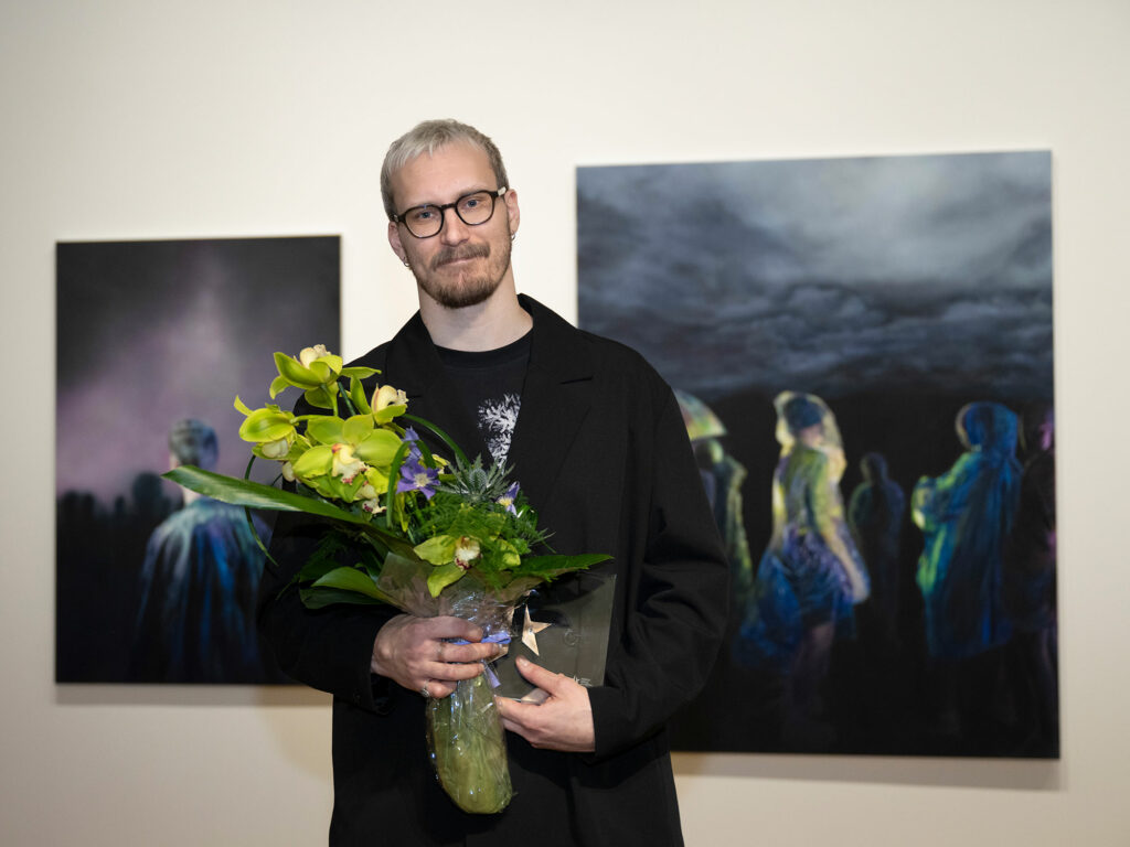 Pitkä vaalea mies seisoo kahden maalauksen edessä kukat kädessään.