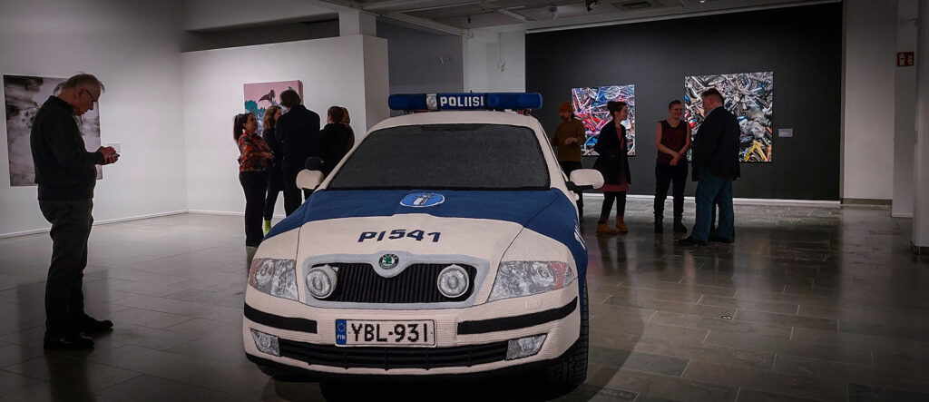 Näyttelytila, jossa on iso virkattu poliisiauto ja muutama ihminen.